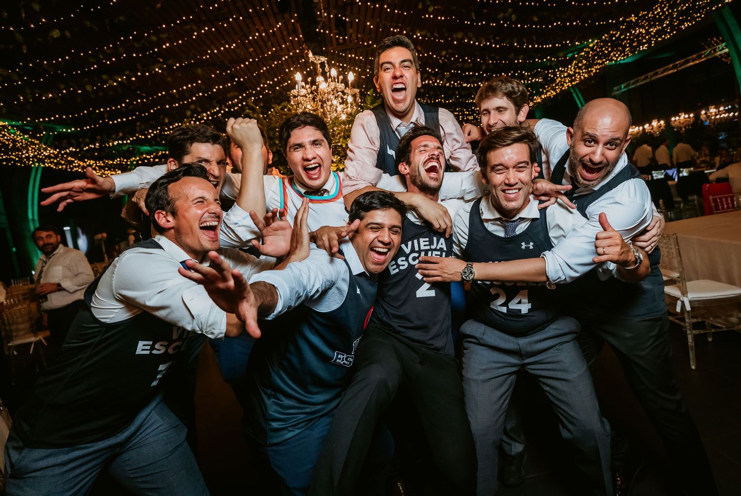 Group of groomsmen in matching jerseys cheering and posing together at wedding reception.
