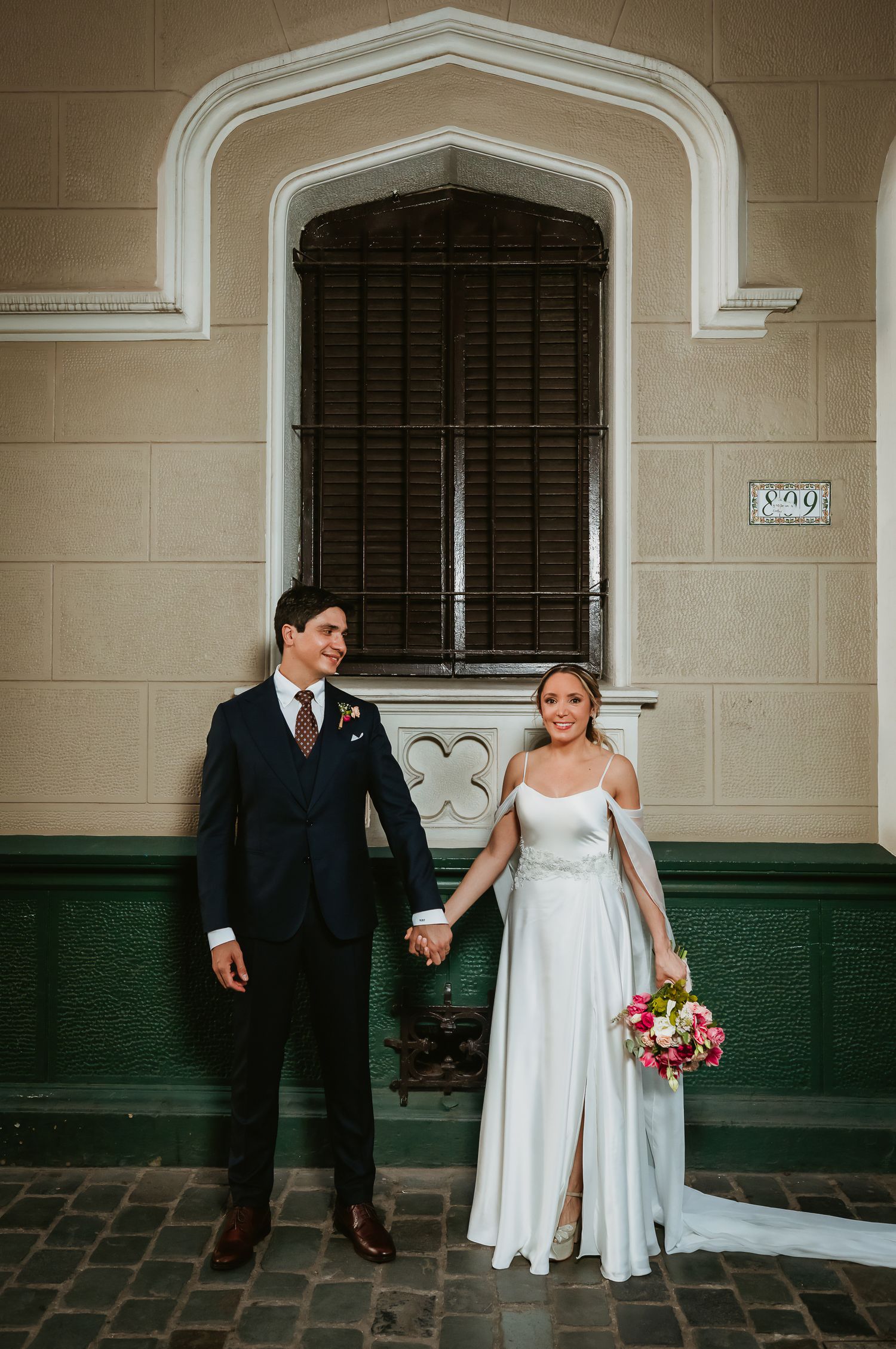 Bride and groom holding hands in front of green and beige building, bride in white gown with bouquet.