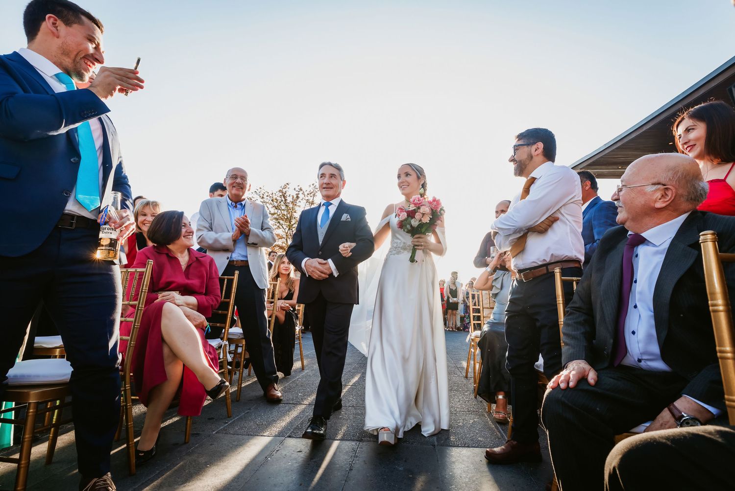 Bride in white gown walks down outdoor aisle escorted by father at sunny wedding ceremony.