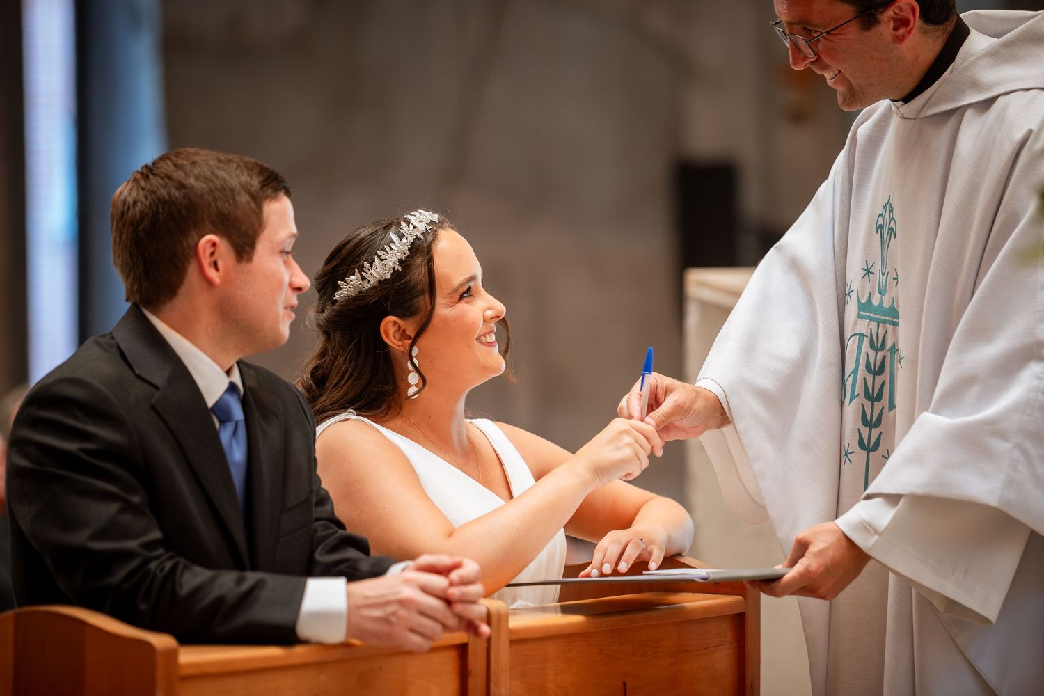 Bride smiles as priest presents the marriage register for signing during Catholic wedding ceremony.