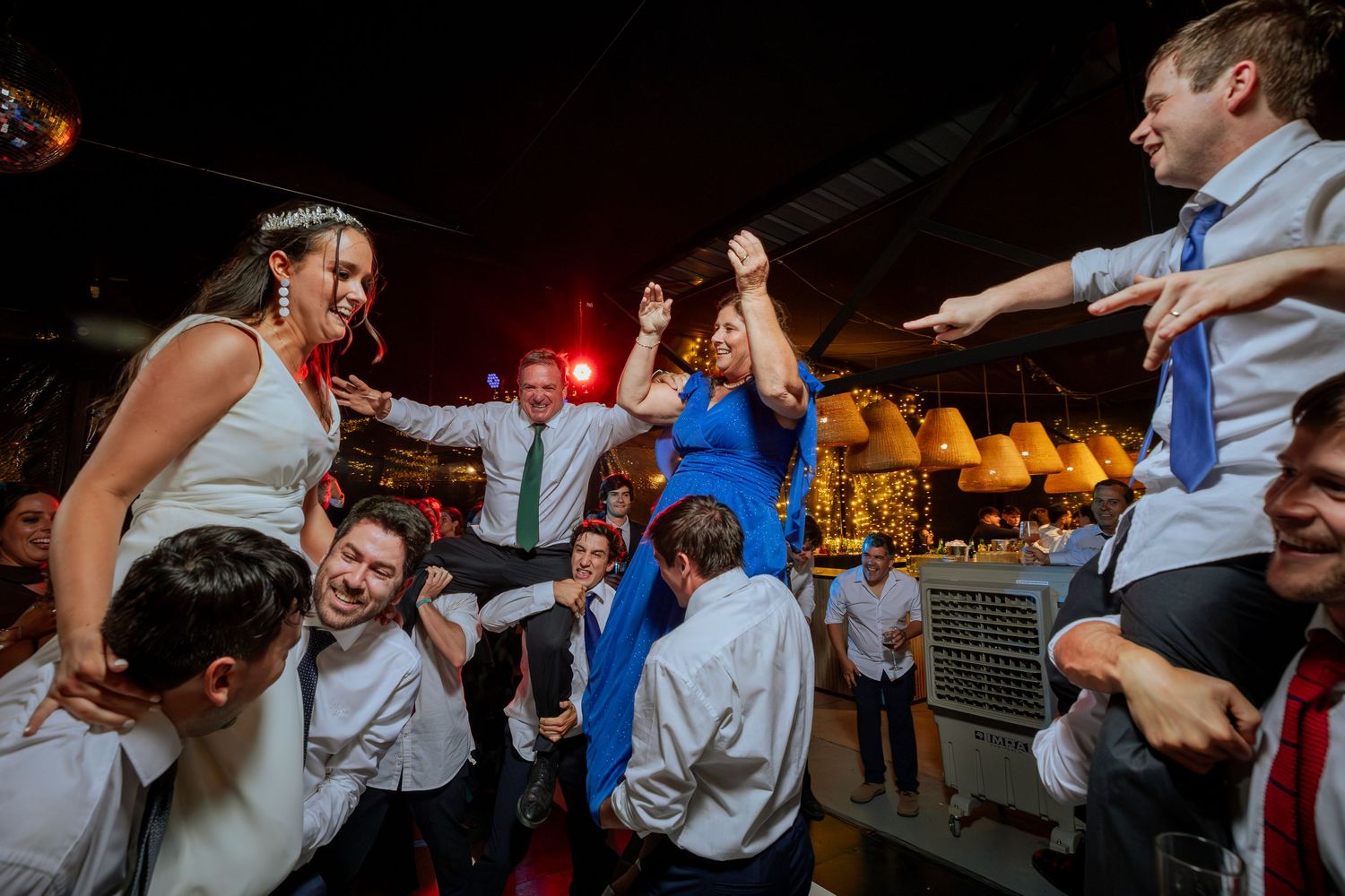 Bride in tiara and guests dancing energetically on a crowded wedding reception dance floor.