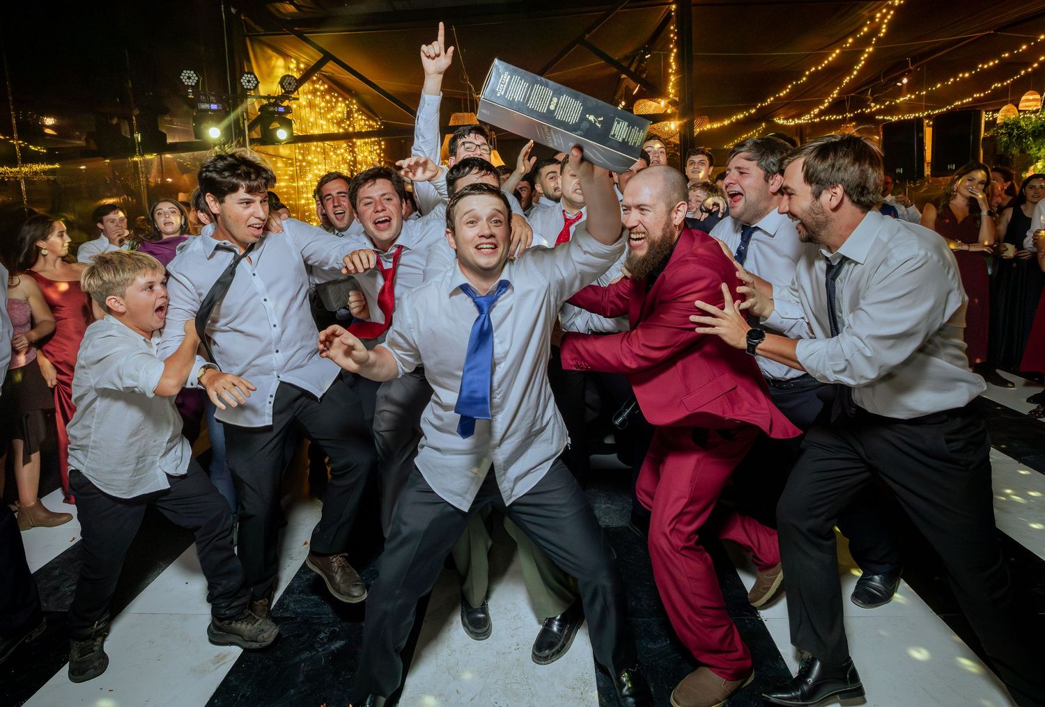 Groom in burgundy suit celebrates with groomsmen on wedding reception dance floor.