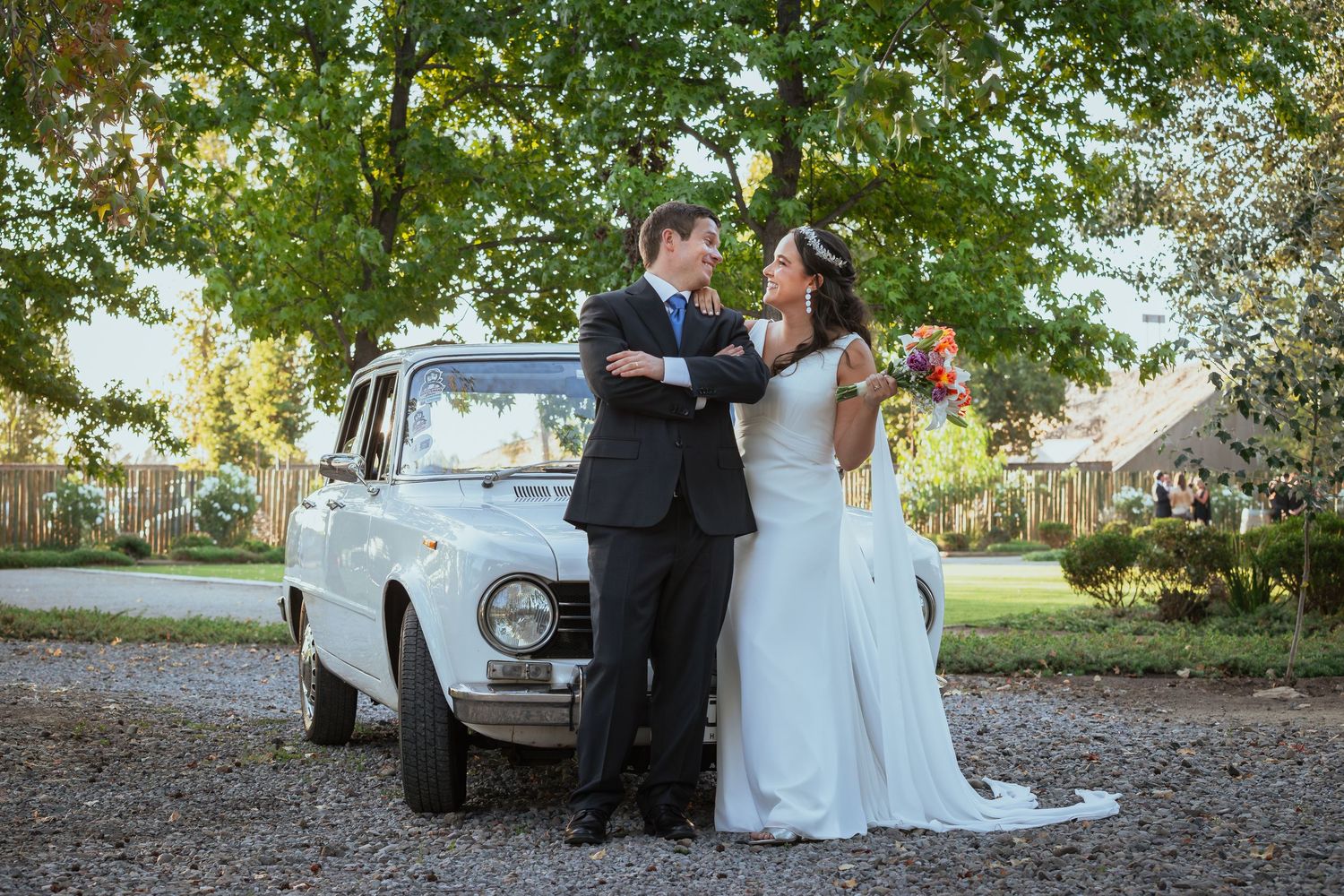 Bride and groom sharing a tender moment beside a classic white vintage car on a gravel path.