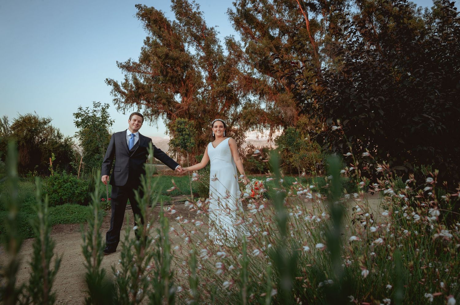 Bride and groom holding hands walking through a lush garden at golden hour on their wedding day.
