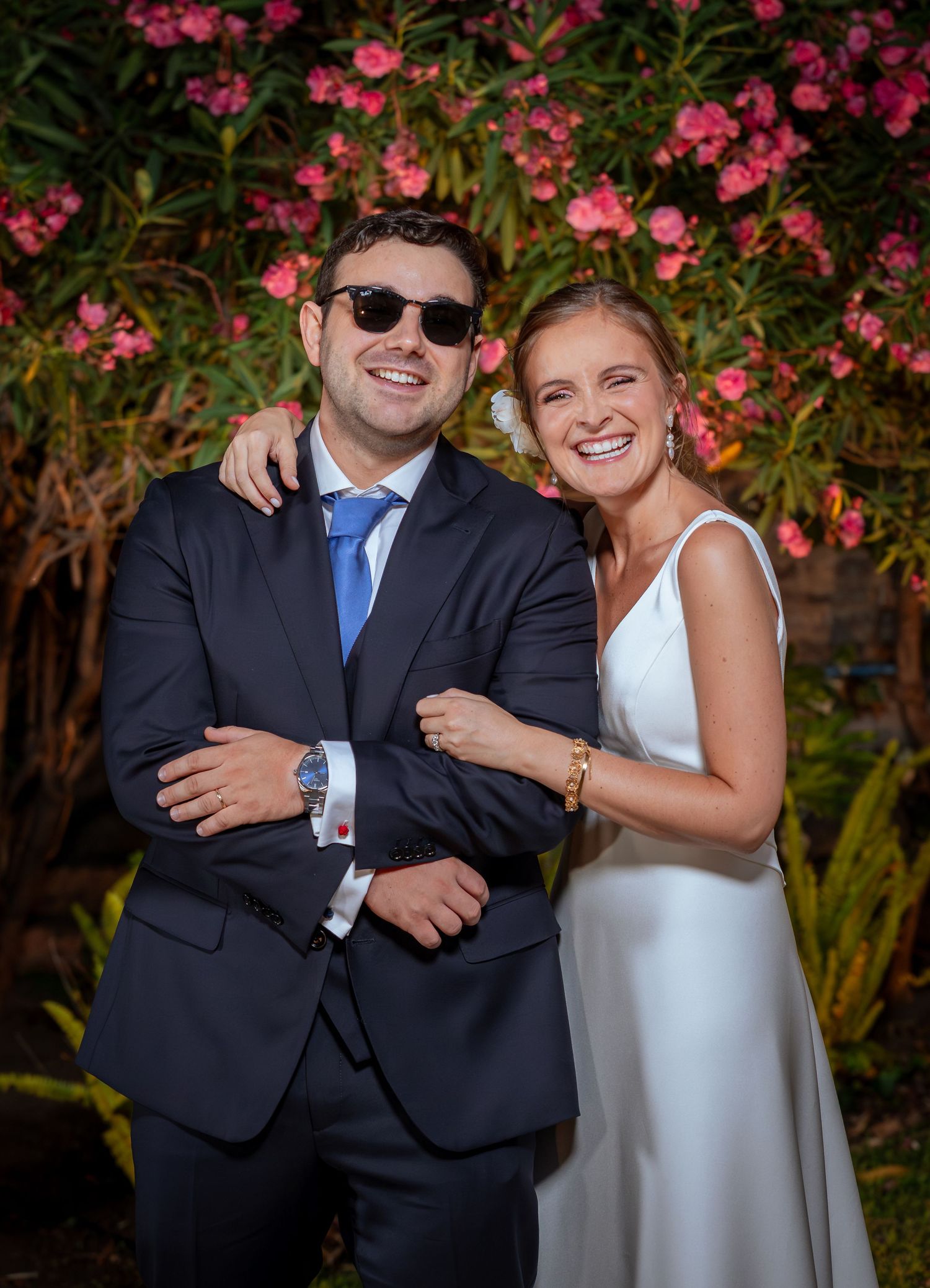 Laughing bride and groom in sunglasses pose playfully before pink flowering bushes at wedding.