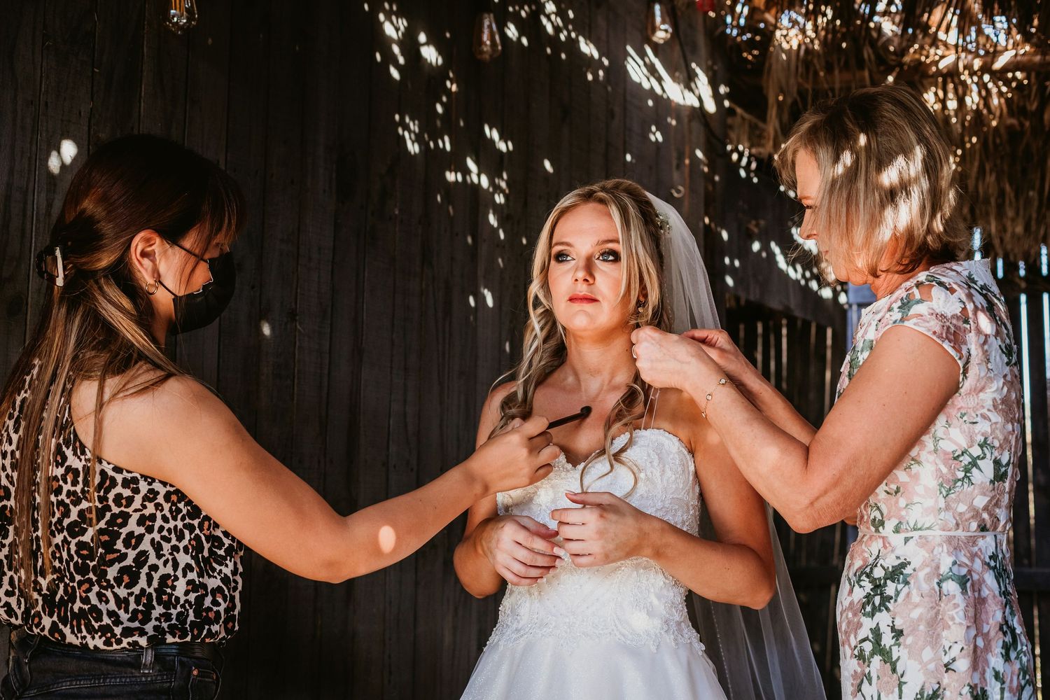 Two women helping bride with final dress preparations inside rustic barn wedding venue.