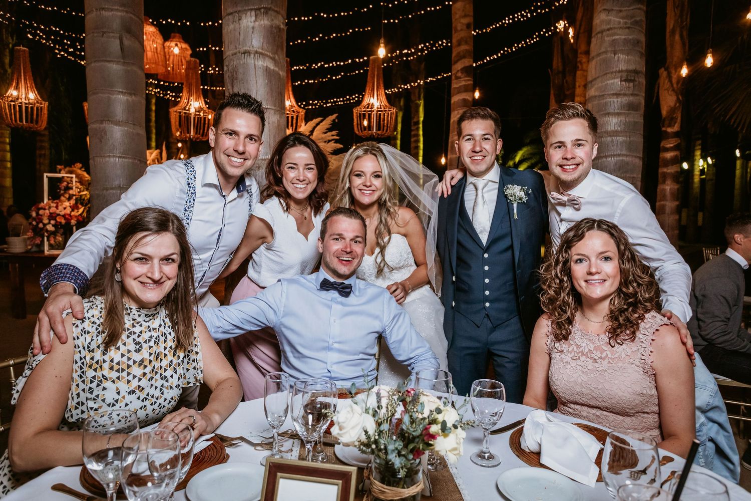 Wedding couple poses with smiling friends and family at elegantly decorated reception table.