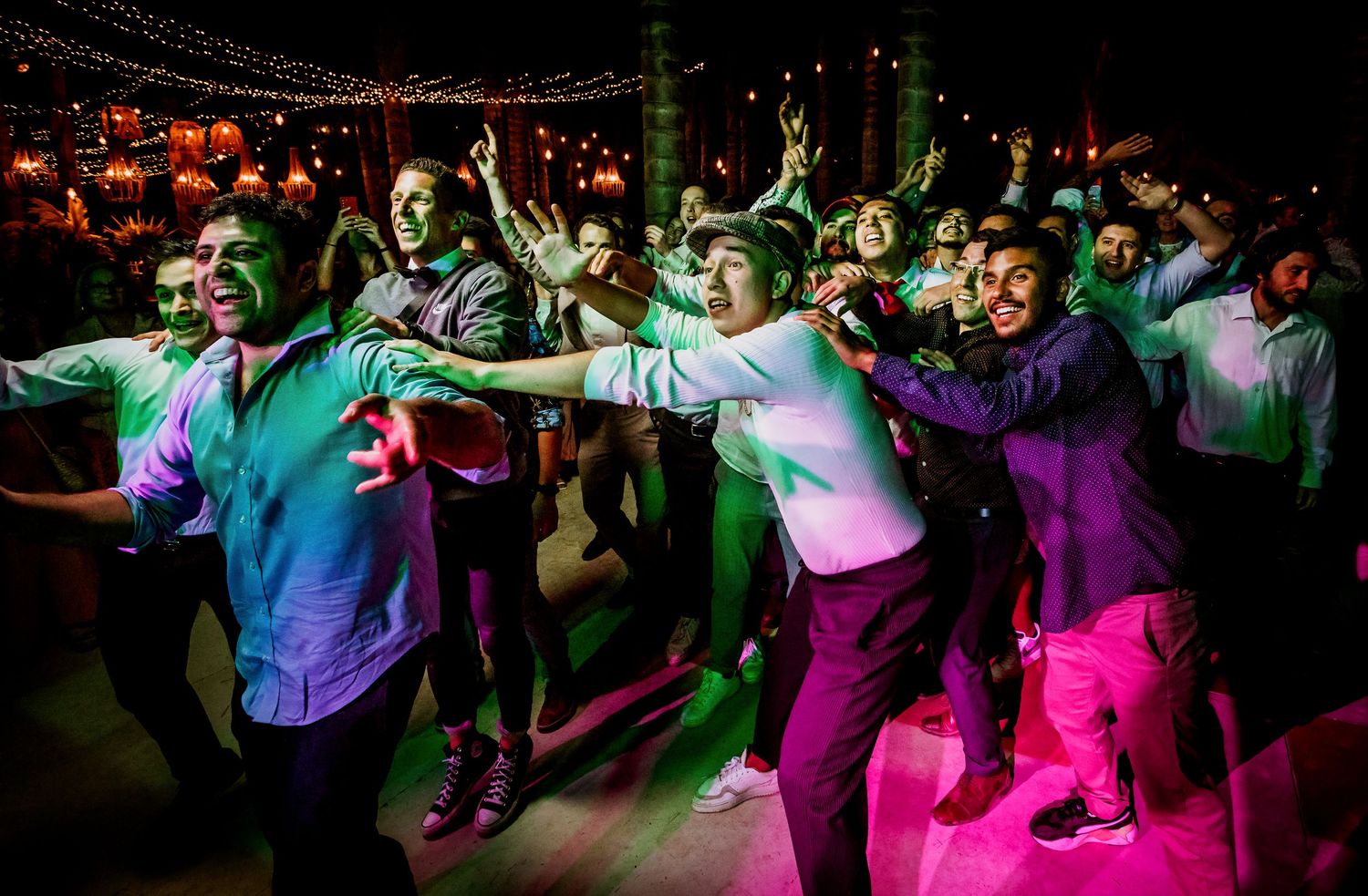 Wedding guests dancing energetically together on the dance floor under colorful lights at reception.