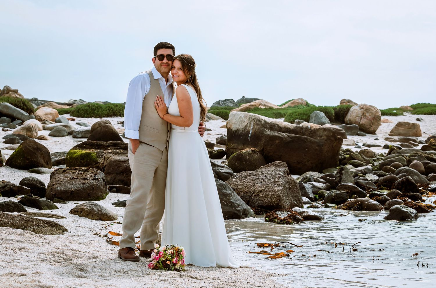 Bride and groom embracing on rocky shoreline with large boulders and calm water behind them.