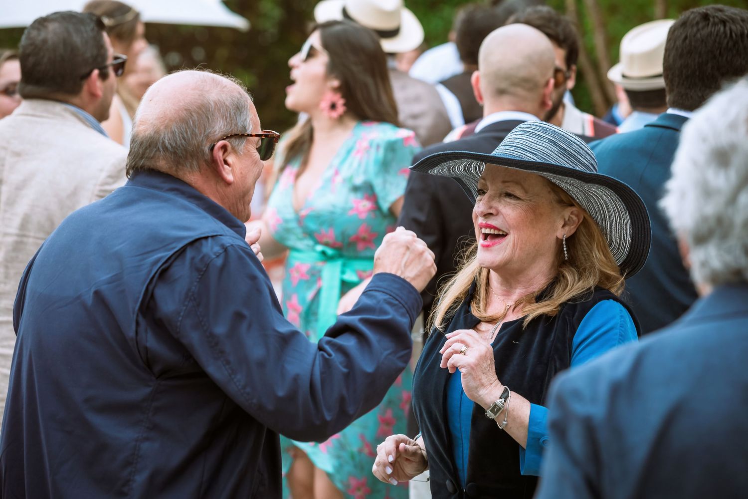 Older couple dancing and laughing at an outdoor wedding, woman wearing a wide-brim hat.