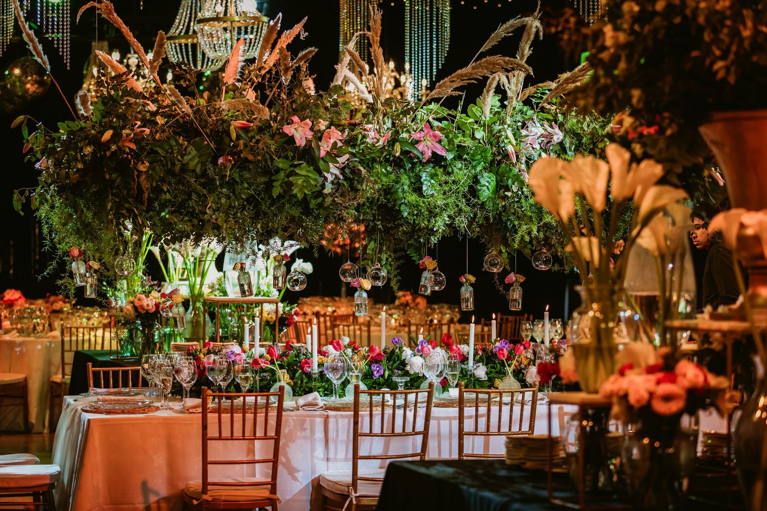 Opulent wedding reception table decorated with towering lush floral arrangements and warm candlelight.