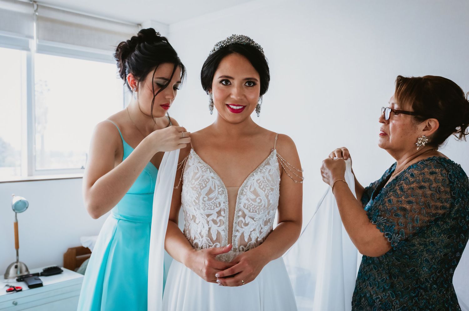 Bride in white lace gown being helped by bridesmaid and mother while getting ready for wedding ceremony.