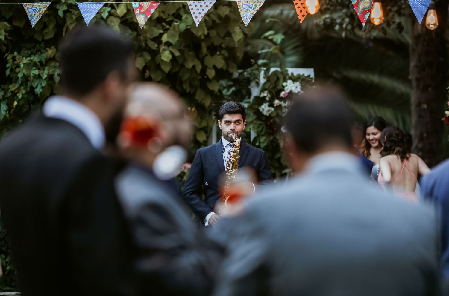 A man in a navy suit stands at an outdoor wedding ceremony decorated with colorful bunting.