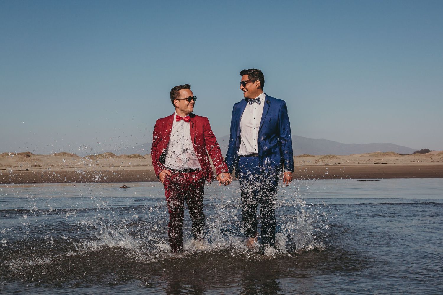 Two grooms in red and blue suits walk hand-in-hand through shallow water under a clear blue sky.