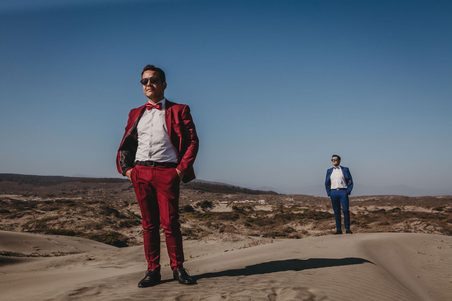 Two grooms in red and blue suits posing on sandy desert dunes under clear blue sky.