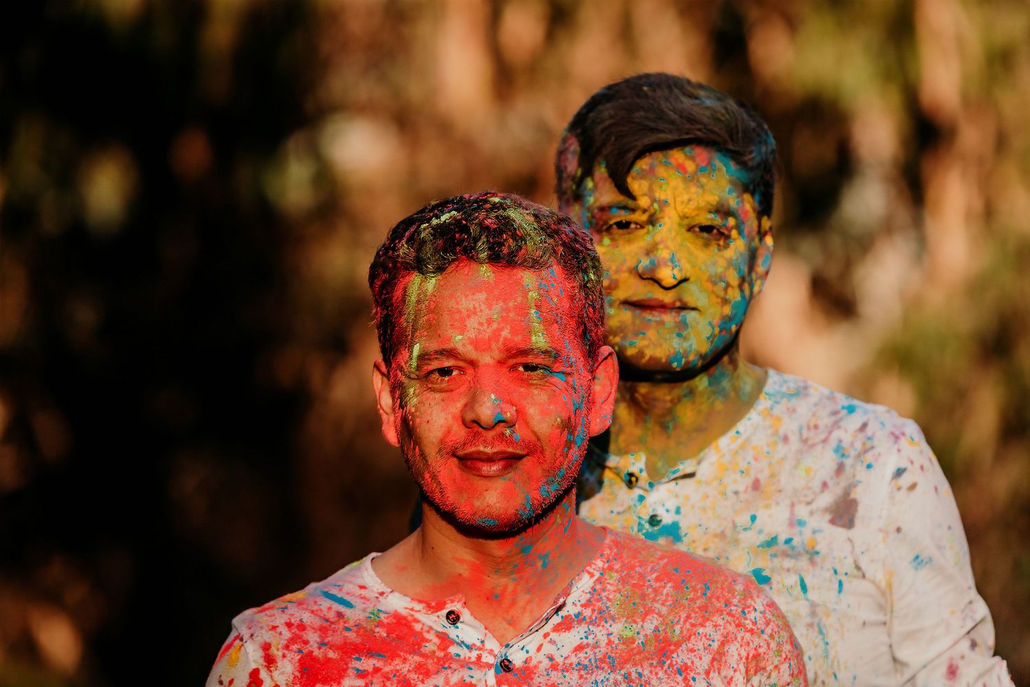 Two men smiling, faces covered in vibrant multicolored Holi powder paint in an outdoor setting.