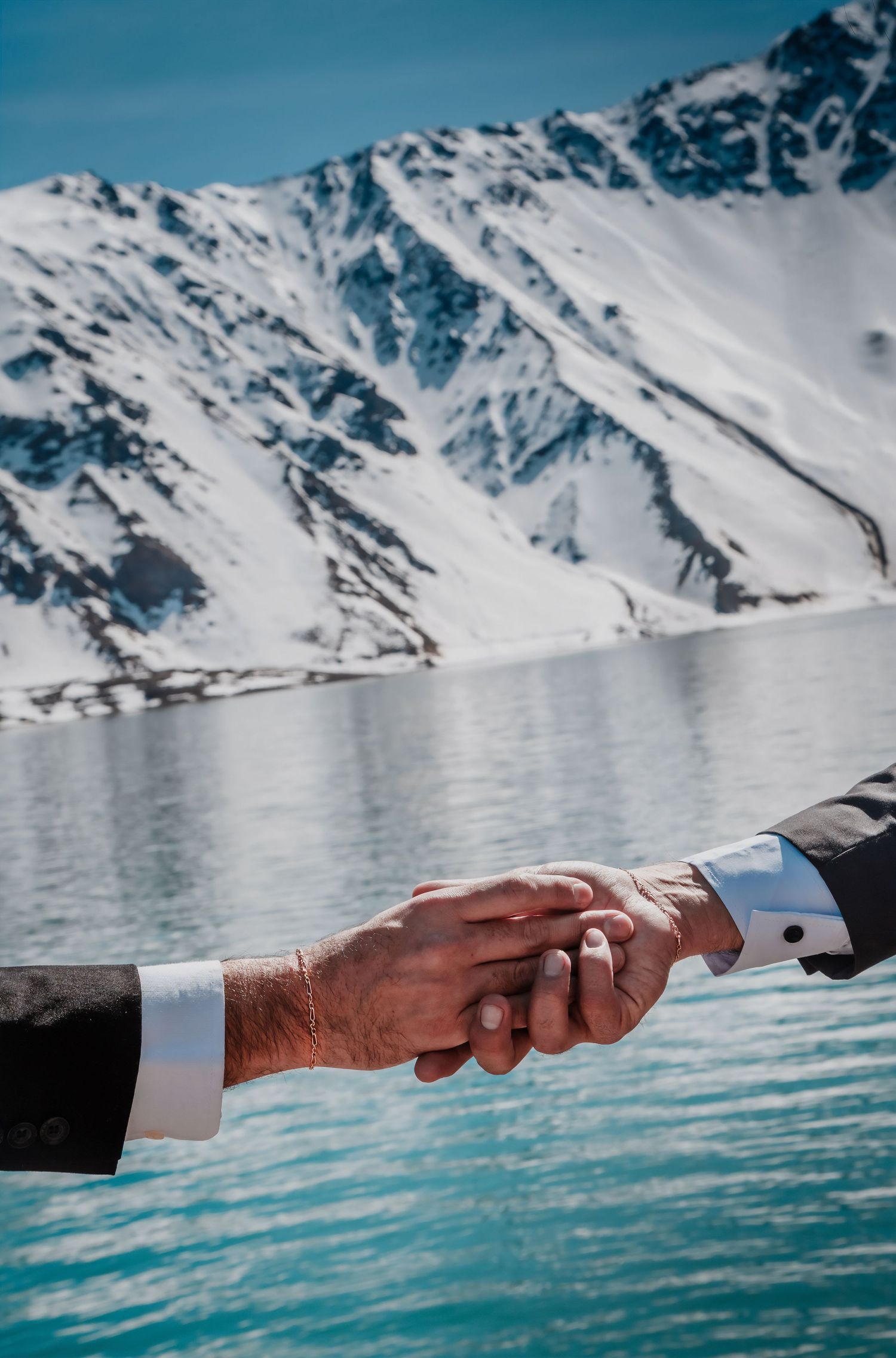 Close-up of two grooms holding hands with a snowy mountain lake in the background.