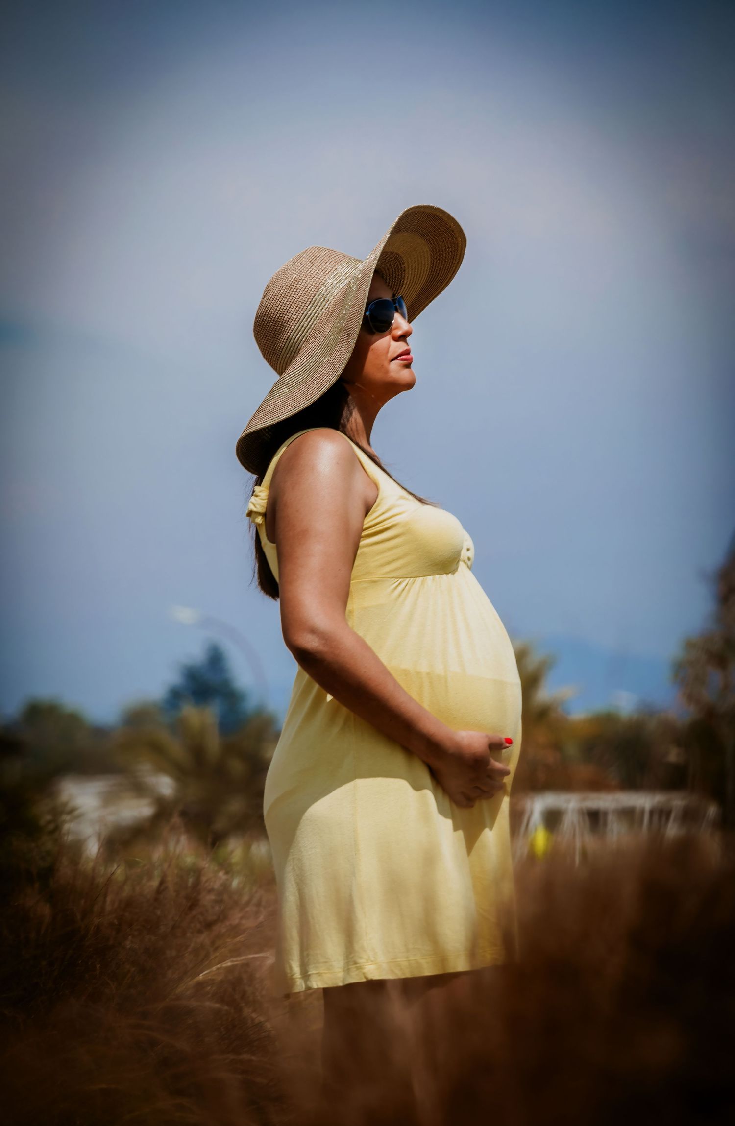 Pregnant woman in yellow dress and wide-brim hat cradling her belly outdoors in a sunny field.
