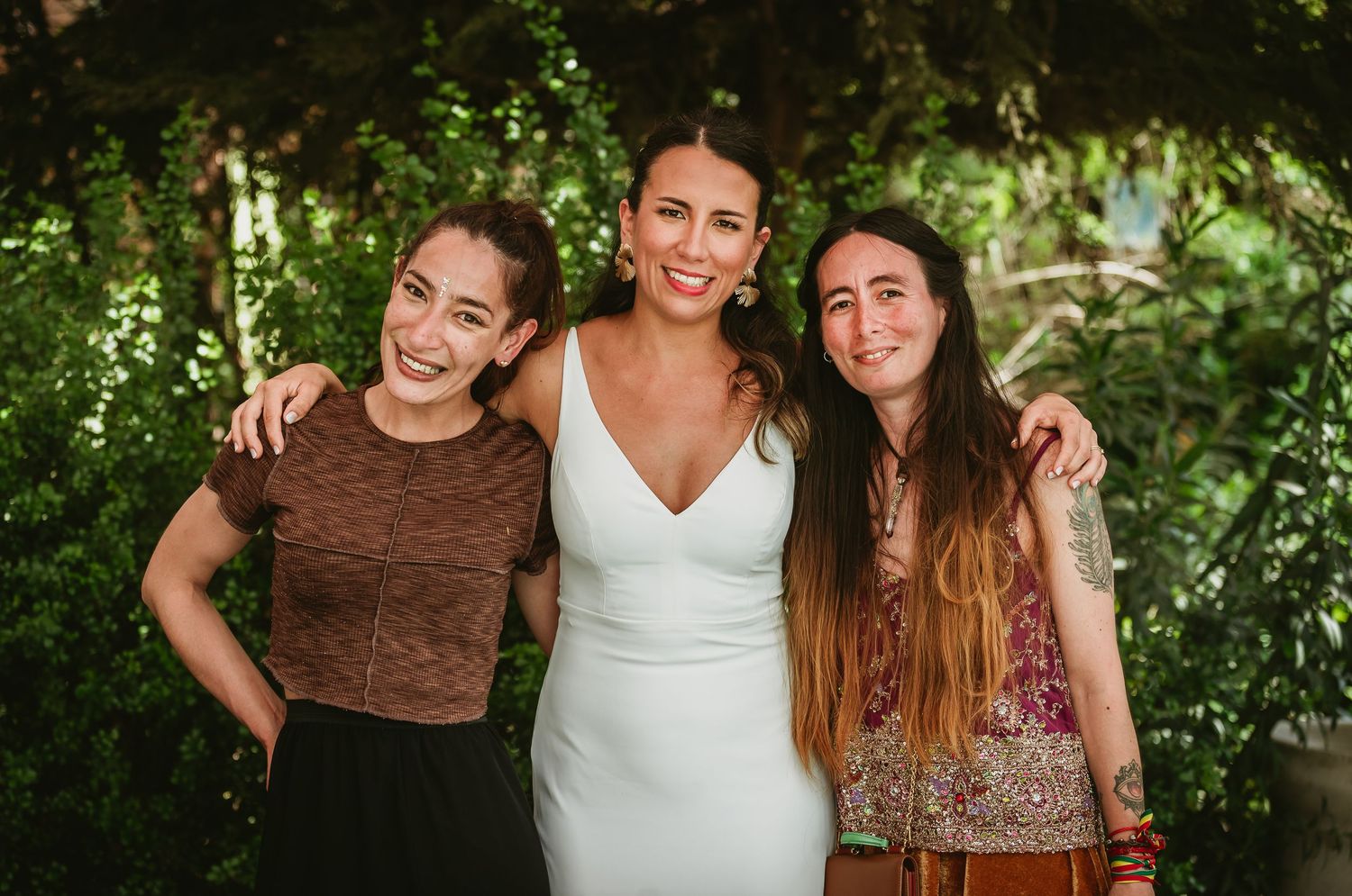 Three women smiling together outdoors, middle woman in white dress, flanked by friends in brown top and floral outfit.