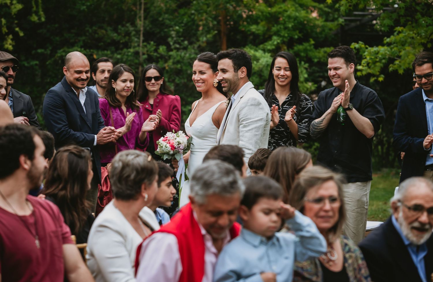 Newlywed couple smiling and walking down the aisle surrounded by clapping wedding guests in an outdoor garden ceremony.