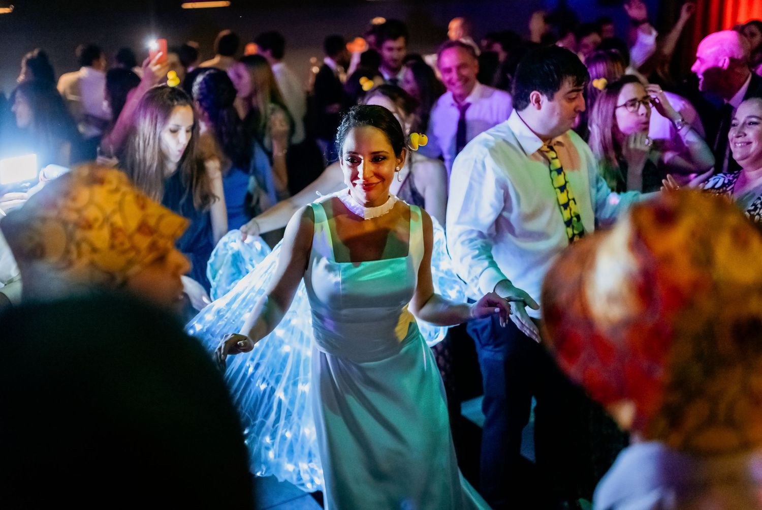 Bride in white gown dancing joyfully on a crowded wedding reception dance floor with colorful lights.