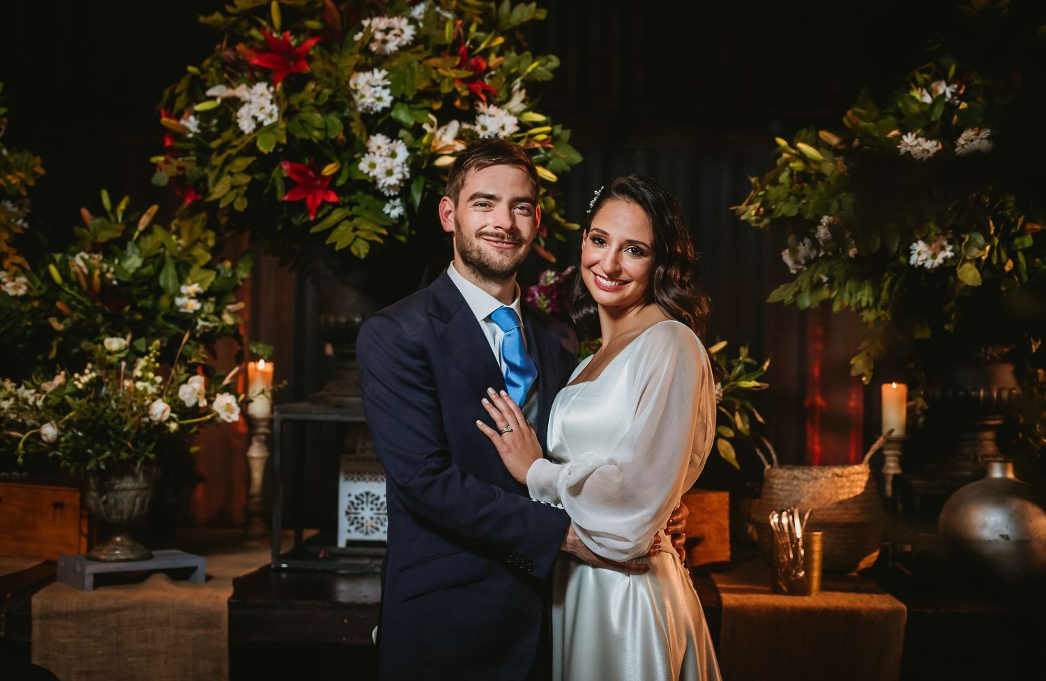 Smiling bride and groom pose together at floral wedding altar with red and white flowers and candles.