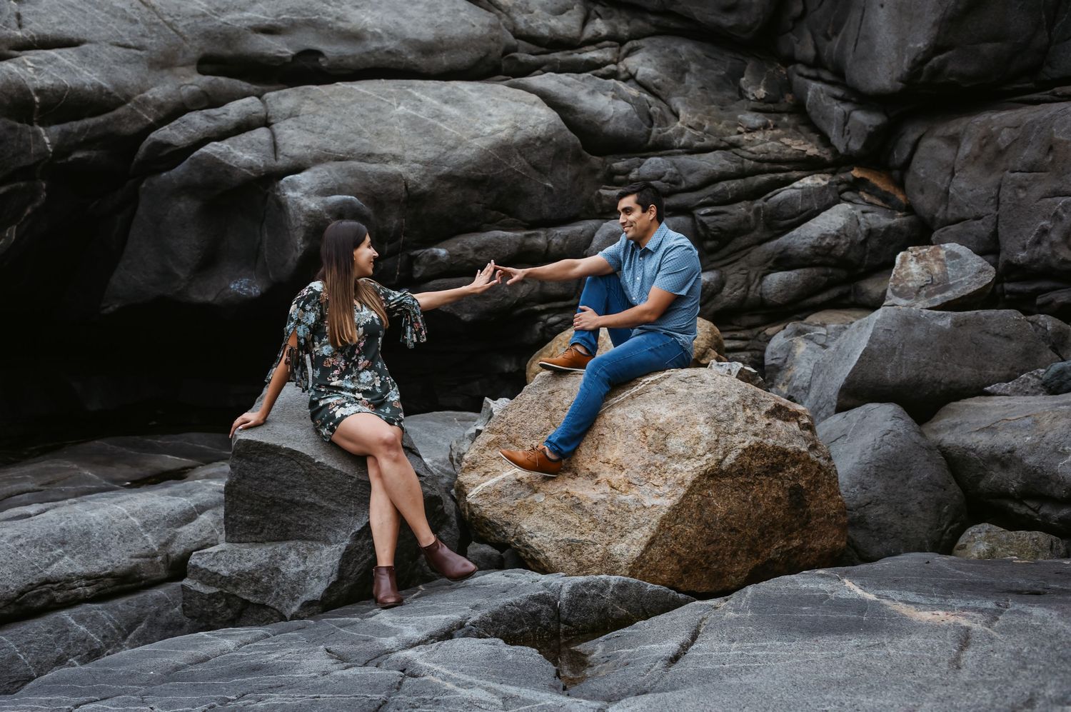 Couple sitting on rocks reaching toward each other in a dramatic rocky coastal landscape.