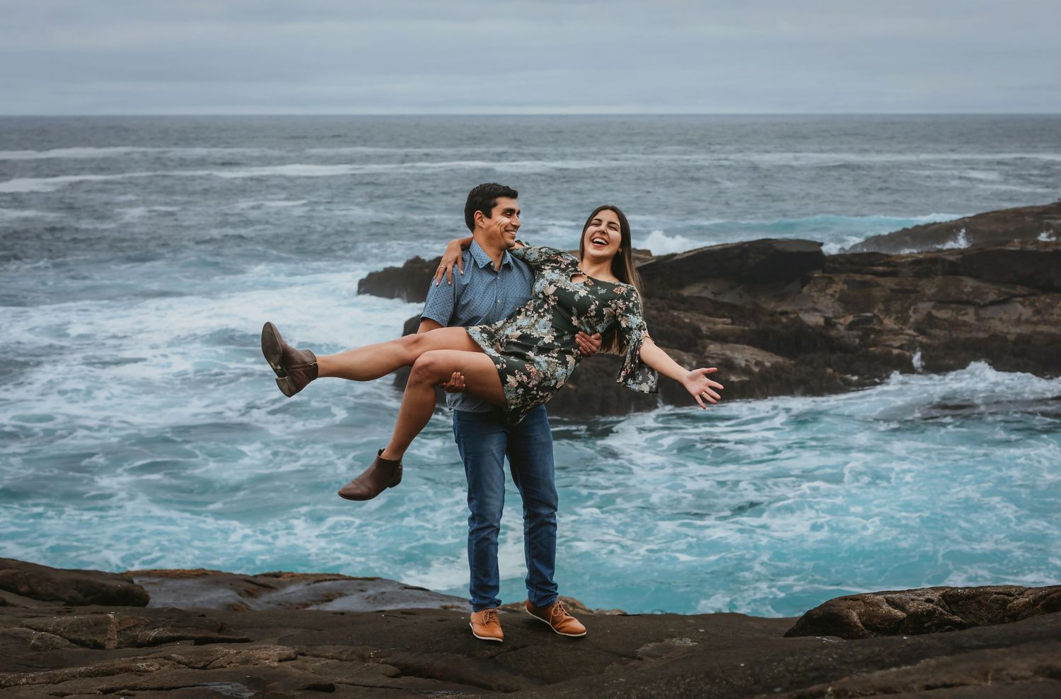 Man carrying woman in his arms laughing on rocky cliff with turquoise ocean waves behind.