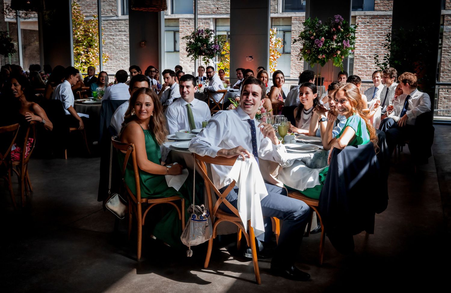 Guests smile and pose at elegantly set wedding reception tables in a sunlit venue.