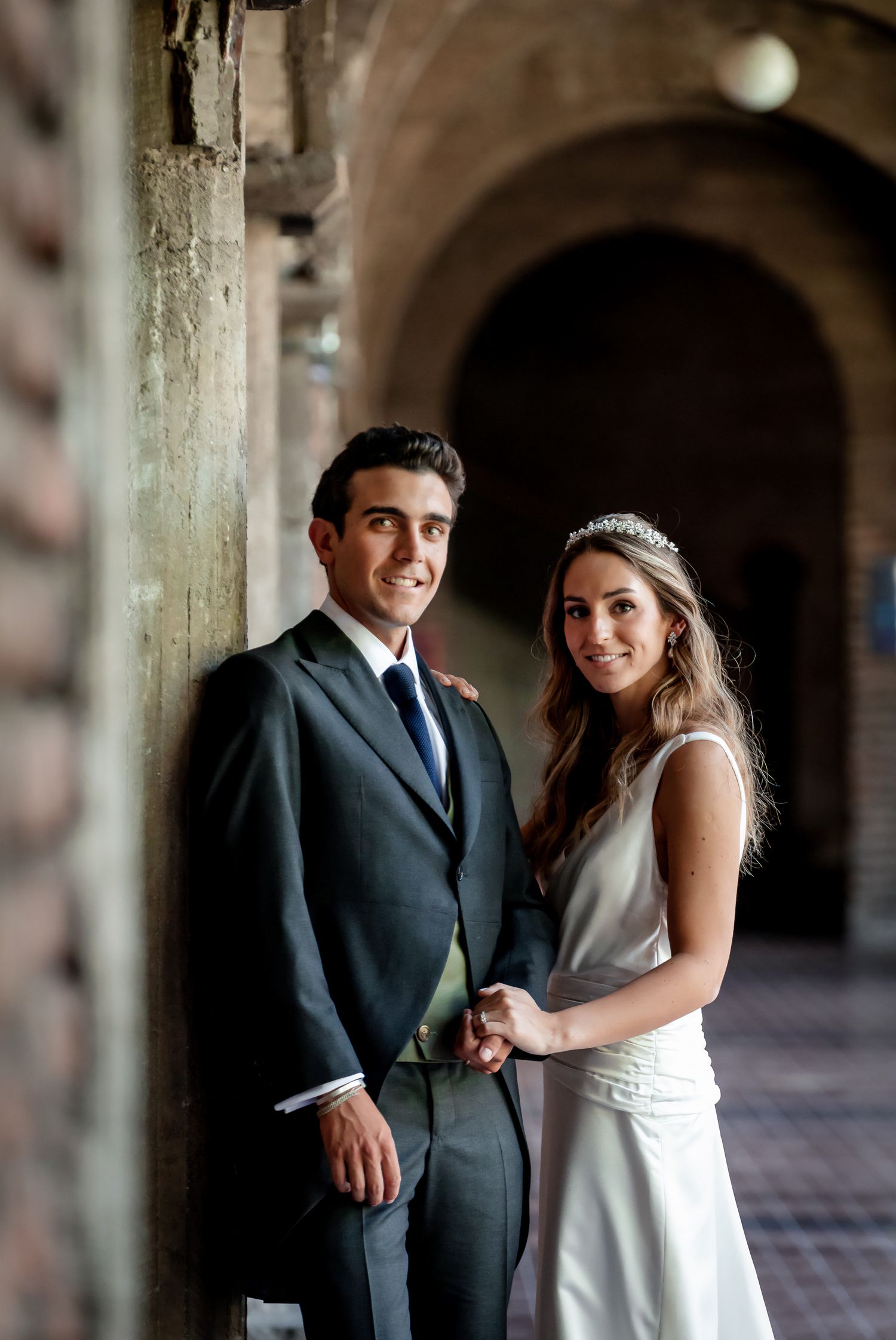Smiling bride and groom holding hands while posing against a stone column in an arched corridor.