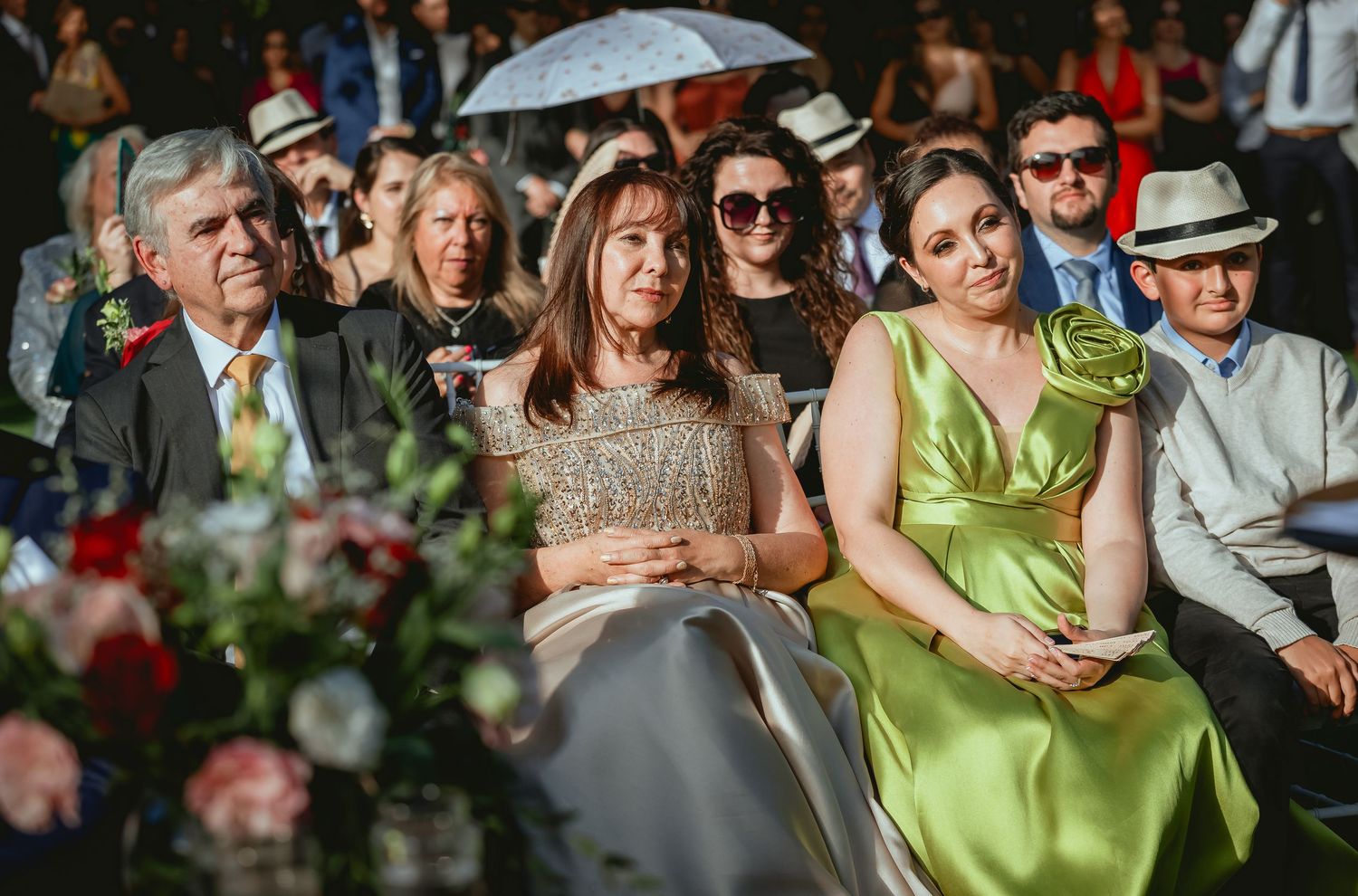 Wedding guests in elegant attire watch outdoor ceremony, woman in green dress seated in front row.