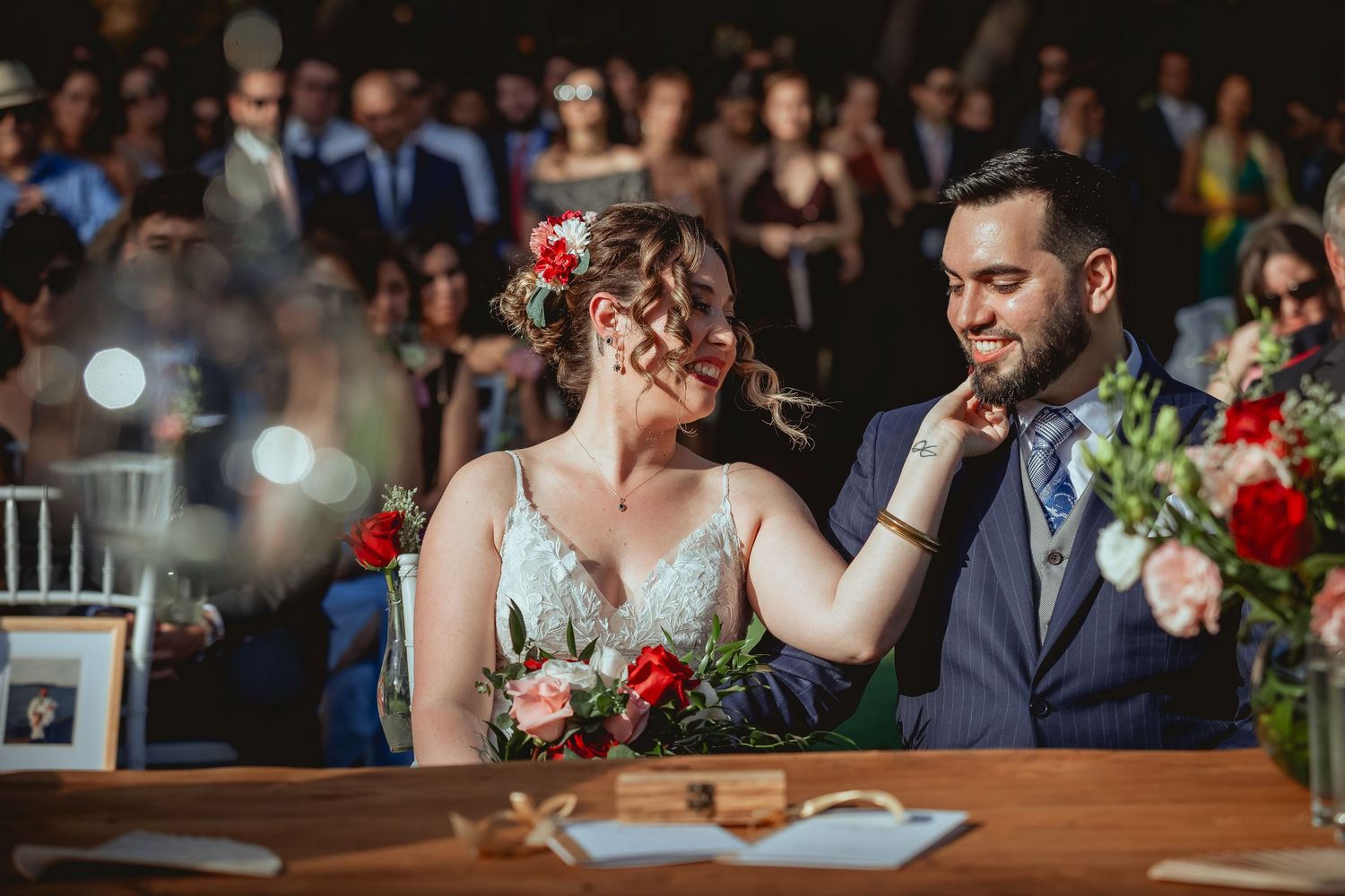 Bride touches grooms cheek tenderly at wedding signing table surrounded by red floral arrangements.