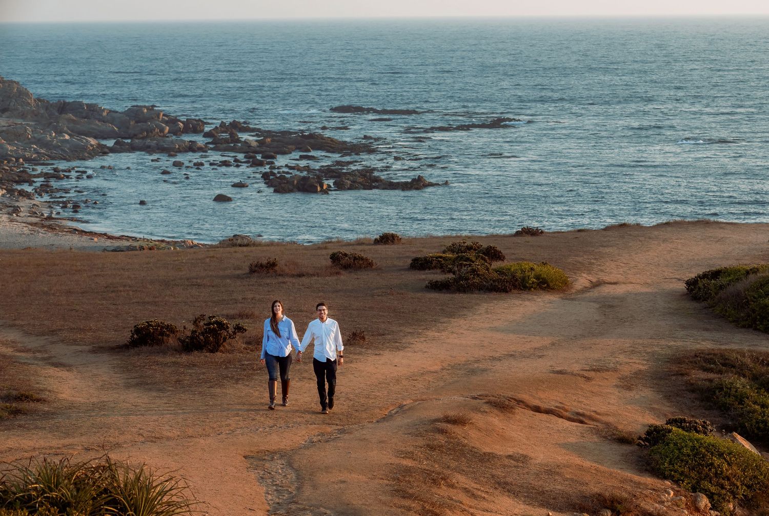 Couple walking hand in hand along a sandy coastal path by the ocean.