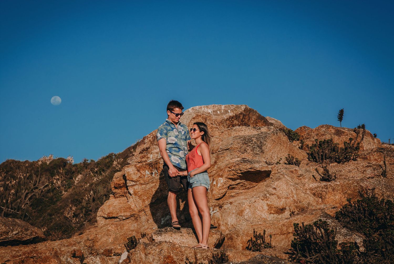 Couple embracing on rocky hilltop under blue sky with visible moon.