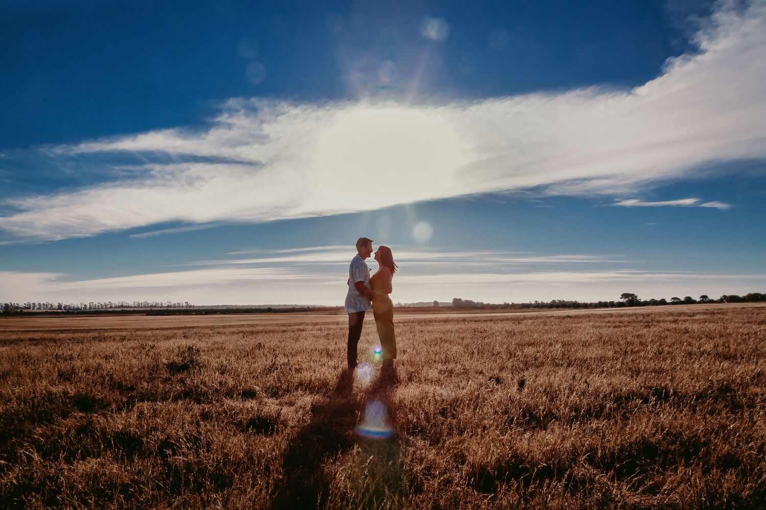 Couple embracing in a sunlit golden field under a blue sky with wispy clouds and lens flare.