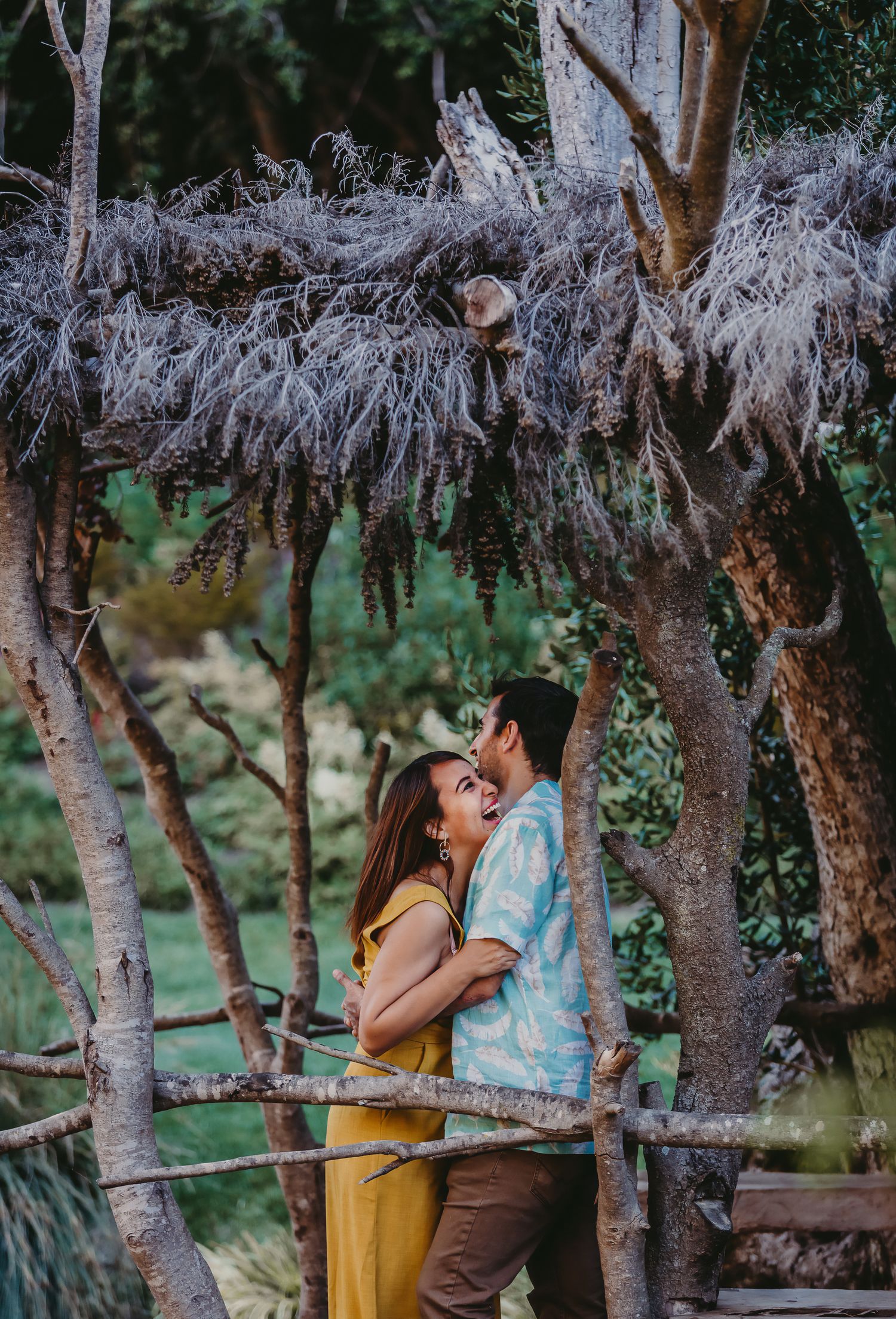 Laughing couple embracing under a rustic wooden arbor draped with Spanish moss in a garden.