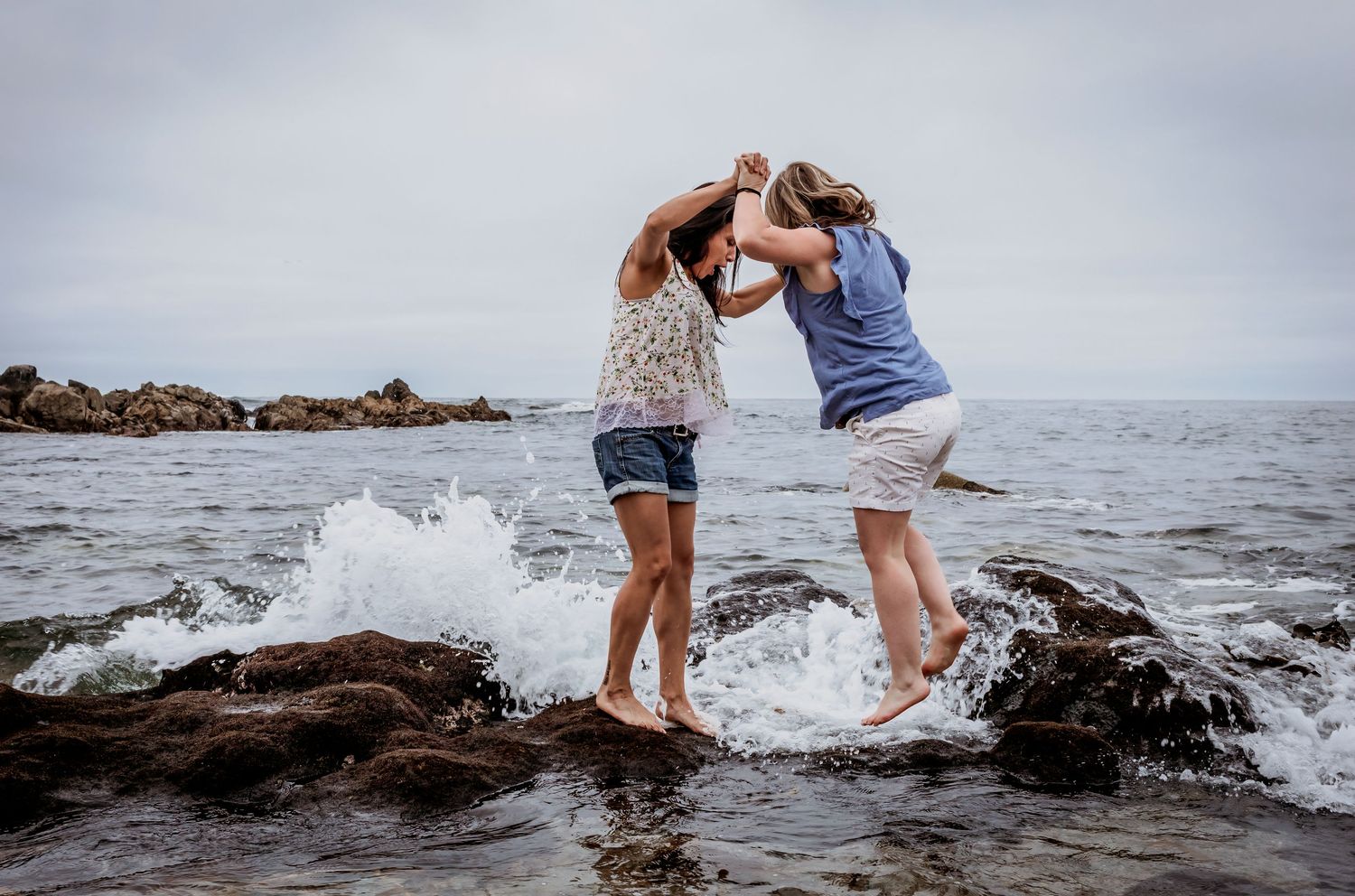Two women playfully spin together on rocky shoreline as ocean waves crash around them.