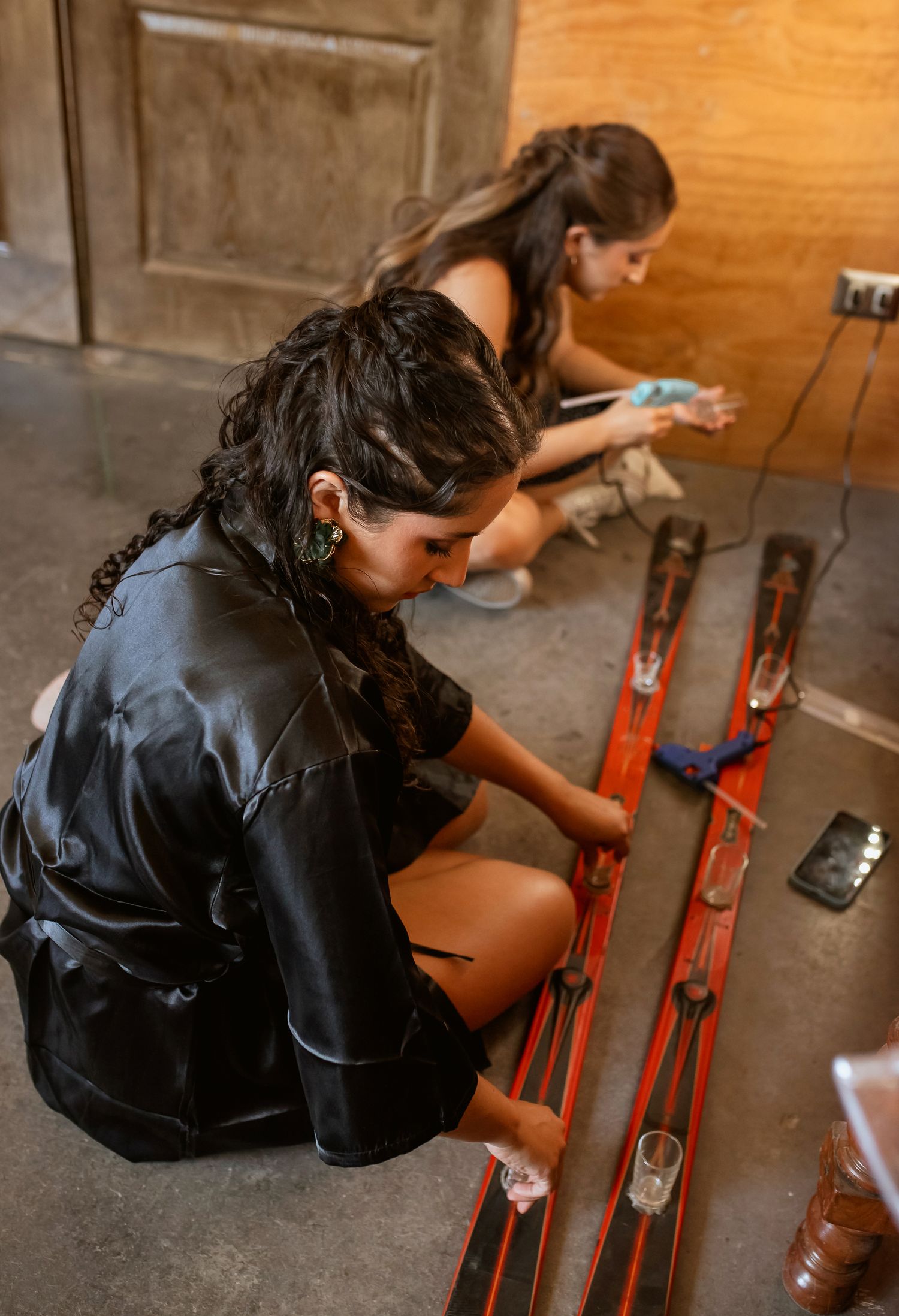 Two women in black satin robes arranging decorative red rails on the floor during wedding preparations.