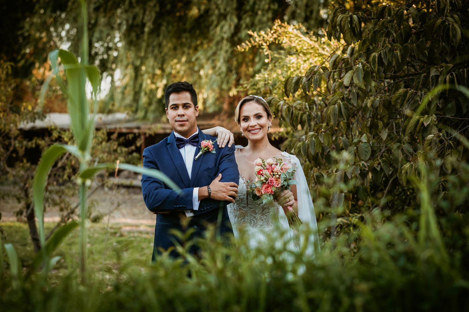 Smiling bride and groom posing together in a lush green garden with colorful bouquet.