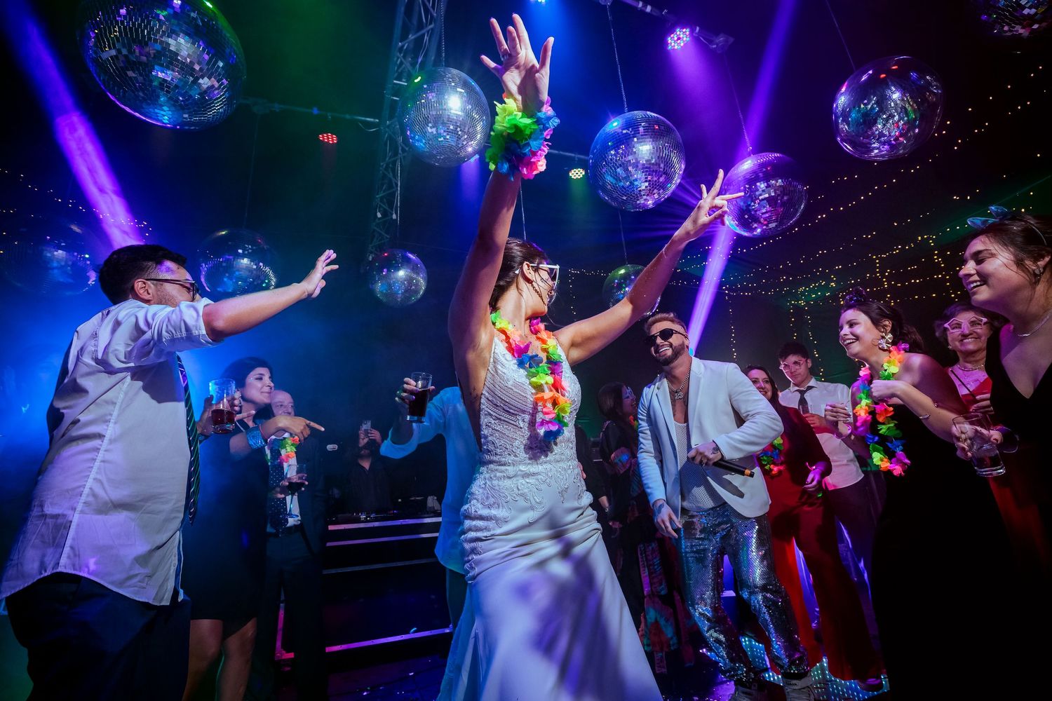 Bride dancing joyfully at wedding reception under disco balls with colorful lights and guests celebrating.