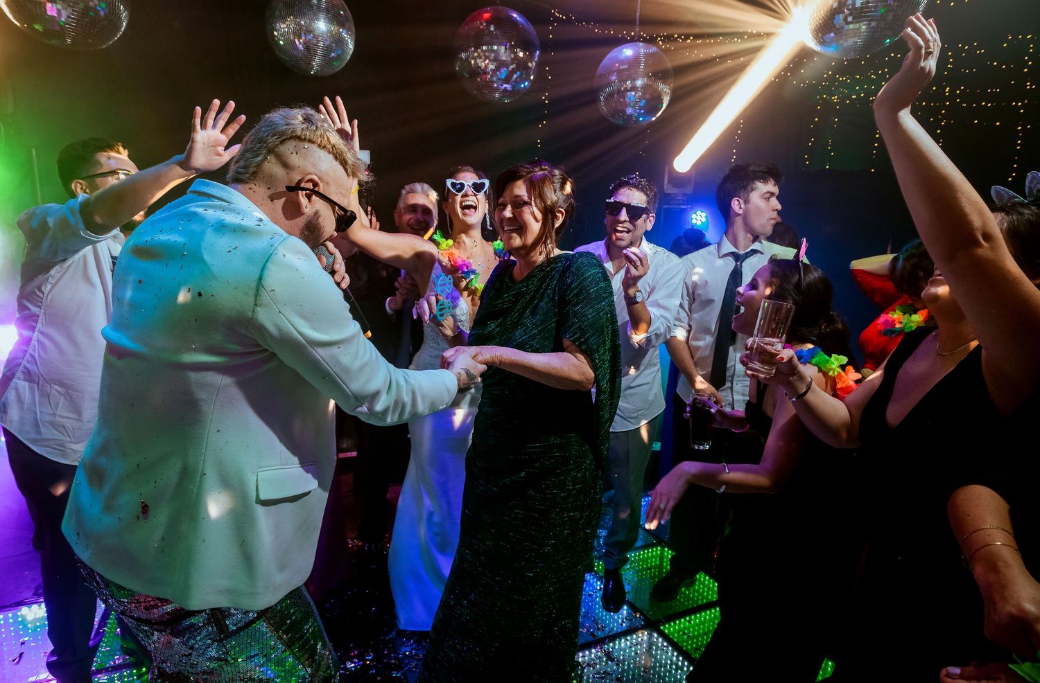 Guests dancing energetically at a disco-themed party with mirror balls and colorful neon lights.