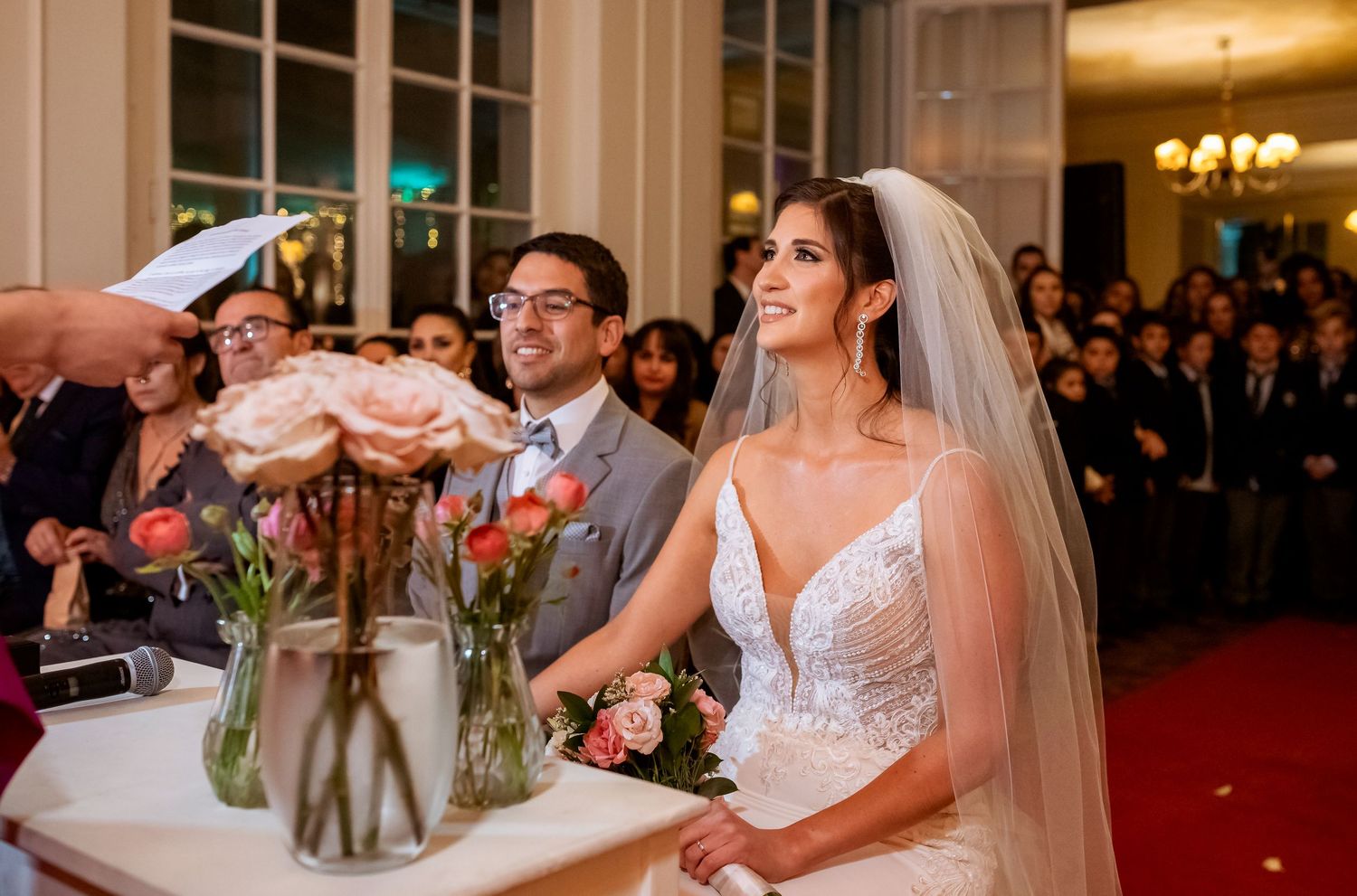 Bride and groom smiling at wedding ceremony altar with pink floral arrangements and guests seated in background.