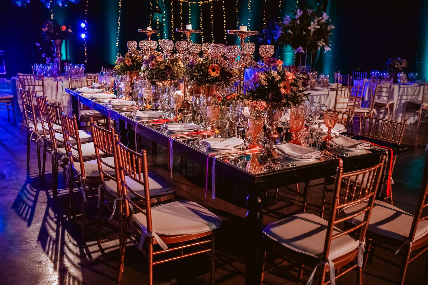 Elegant wedding reception with gold chiavari chairs, long dark tables, floral centerpieces, and ambient blue lighting.