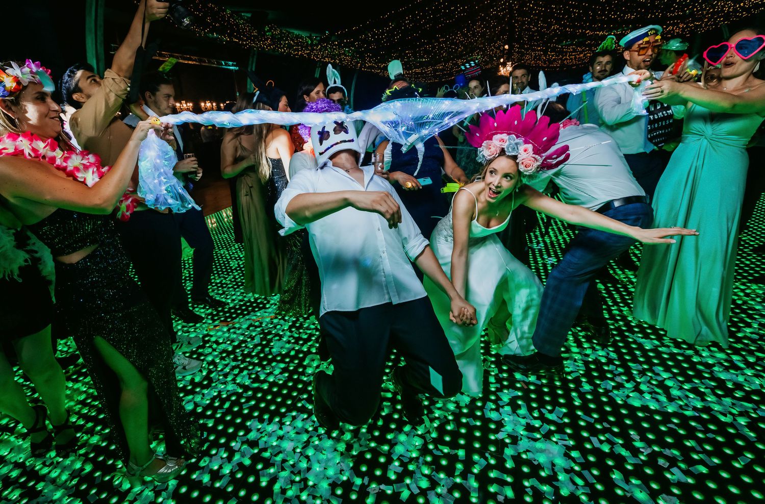 Wedding guests dance under green lights on a sparkling floor while bride in pink crown celebrates joyfully.