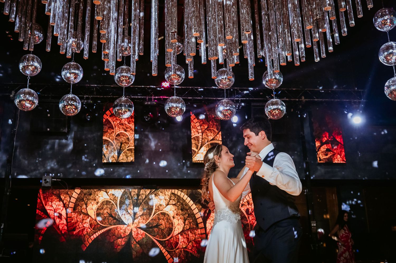 Bride and groom share romantic first dance under disco ball chandelier with vibrant red stained glass backdrop.
