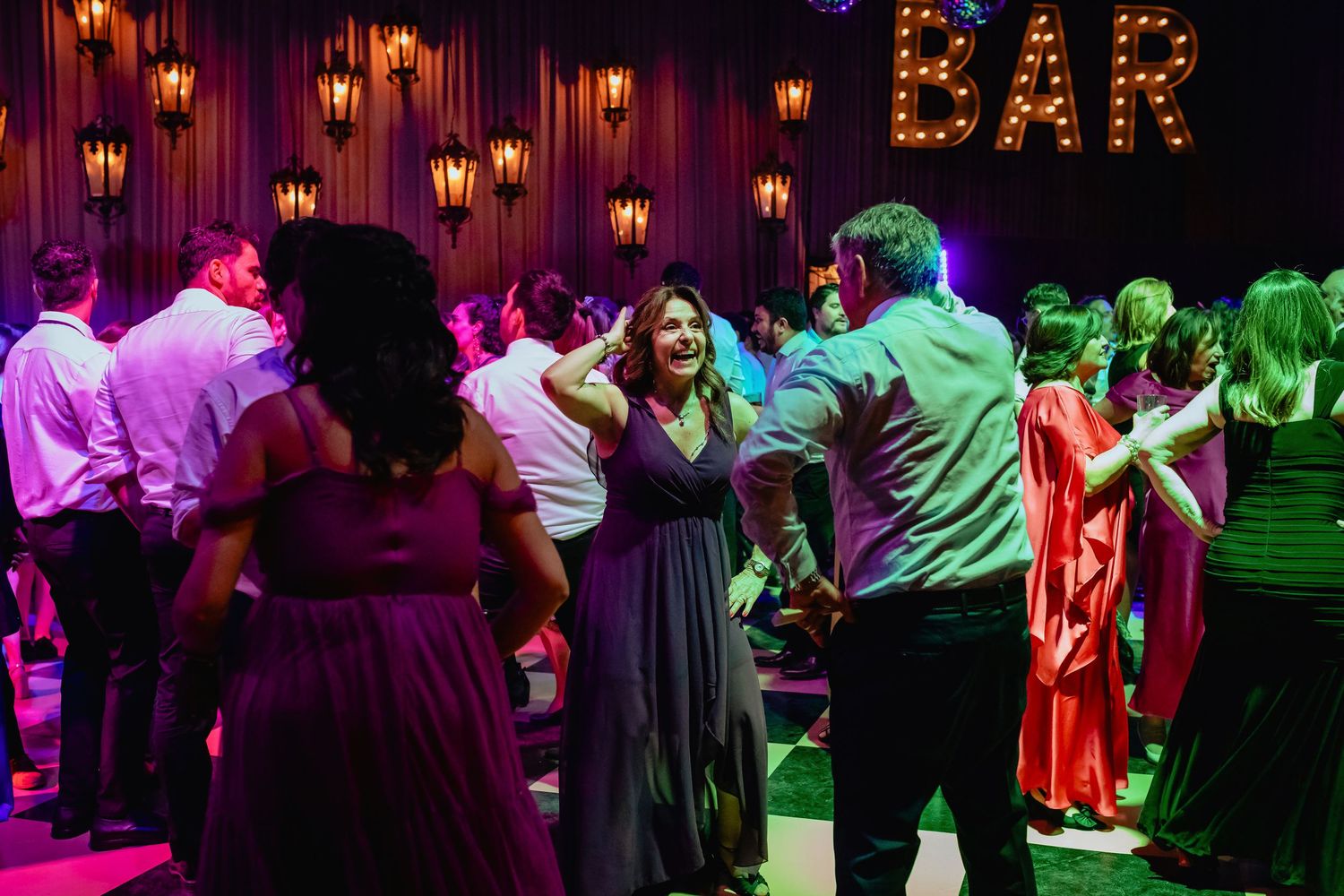 Wedding guests dancing and socializing near a neon BAR sign at a lively evening reception.