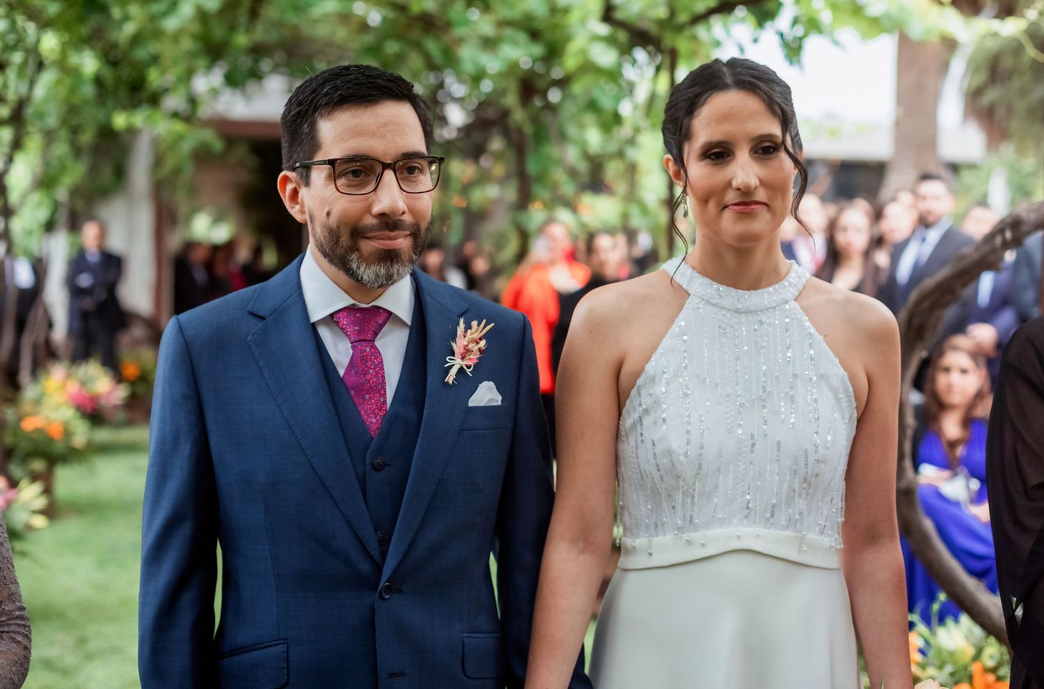 Bride and groom pose together at outdoor wedding ceremony, she in beaded halter gown, he in navy suit.