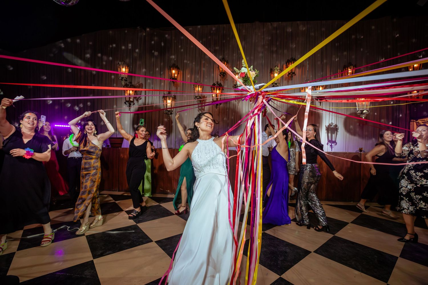 Bride surrounded by guests pulling colorful ribbons on checkered wedding dance floor.