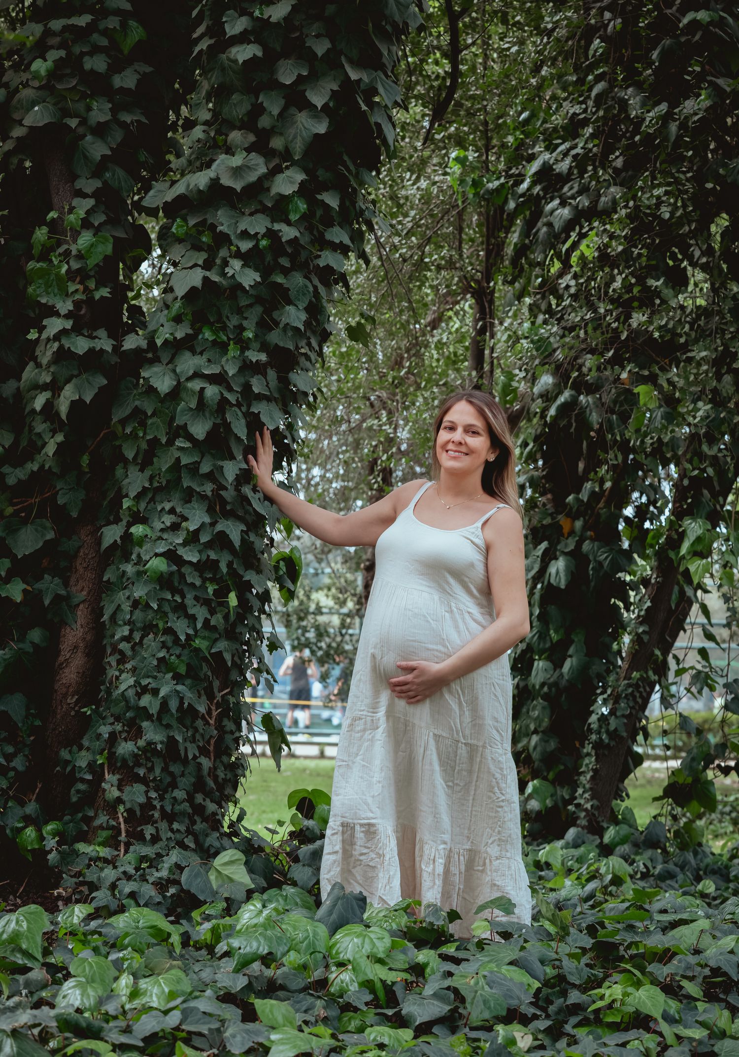 Pregnant woman in white dress standing among lush green ivy-covered trees in a park.