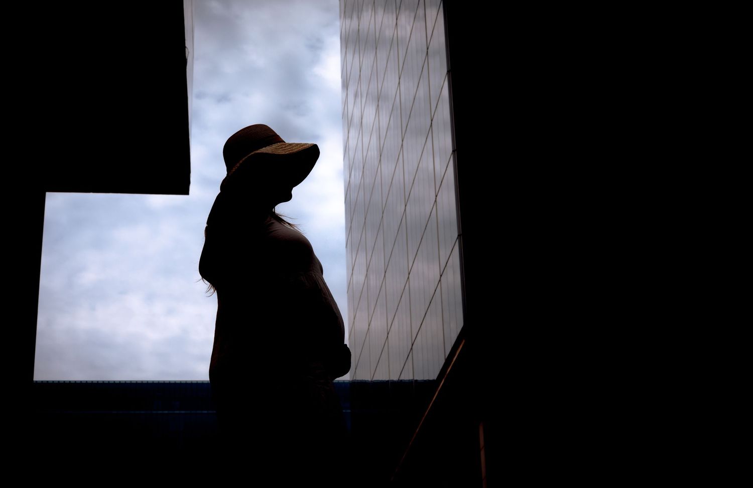 Silhouette of a pregnant woman wearing a hat standing by a window against a dramatic cloudy sky.